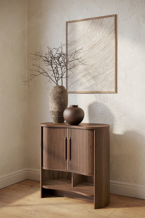 A walnut sideboard with ribbed doors and an open lower shelf stands against a textured beige wall, topped with a decorative stone vase, a rounded brown vase, and a framed piece of textured neutral wall art.