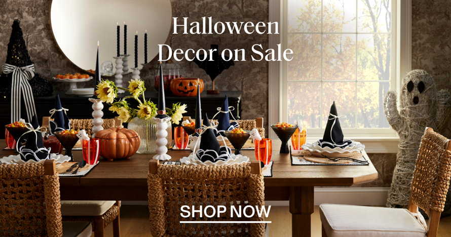 Halloween Decor on Sale. A dining table set for a festive Halloween meal with witch hat place settings, candles, and a spooky ghost figure in the background. Shop now.