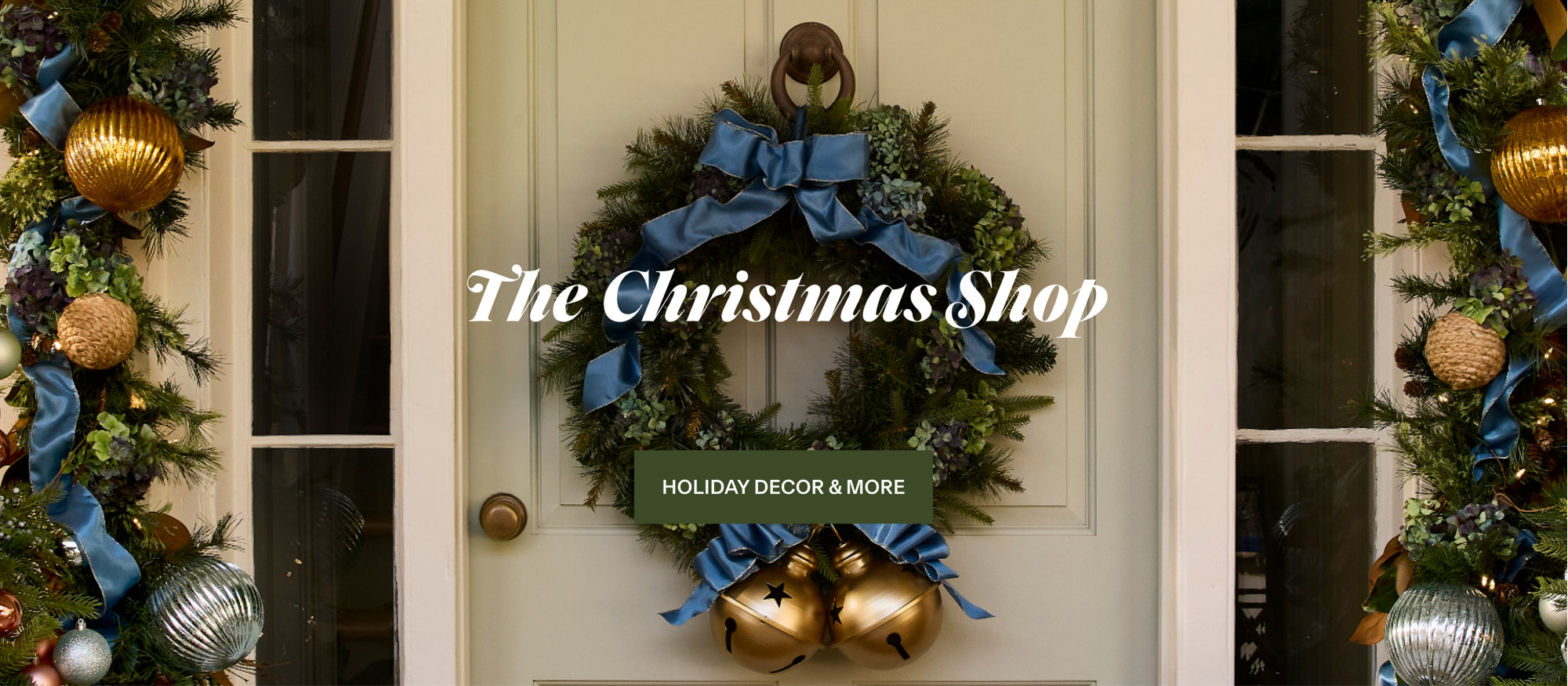 The Christmas Shop, Holiday Decor & More featuring a festive wreath decorated with a blue ribbon and large gold bells hanging on a front door.