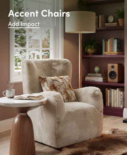 Accent Chairs: Add Impact. A plush, cream-coloured faux fur accent chair sits beside a white pedestal side table with an open book and a steaming mug, set in a cosy corner with a bookshelf and floor lamp.