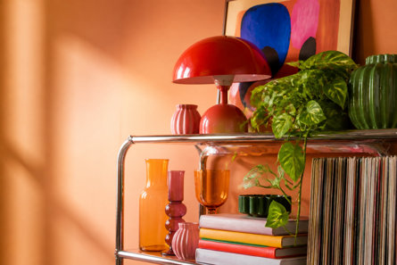 Modern chrome shelving unit styled against a terracotta wall, featuring a bright red mushroom lamp, colourful glass vases, a cascading houseplant, and a stack of vibrant books next to a collection of vinyl records.