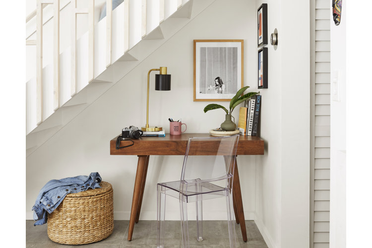 A small wood desk and a clear acrylic office chair set up in an alcove beneath a set of stairs.