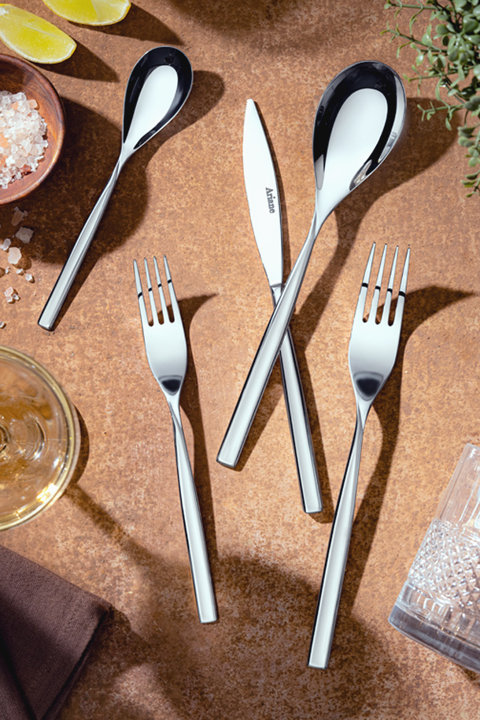 Polished stainless steel flatware set featuring a knife, forks, and spoons arranged on a textured brown surface near lime wedges and glassware.