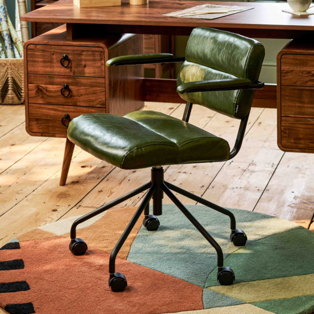 Olive green faux leather office chair with a black metal frame and wheels, placed on a colourful patterned rug in a home office with a wooden desk.