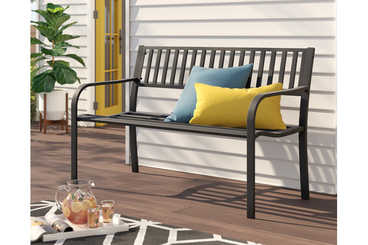 metal outdoor bench with yellow and blue outdoor pillows. Brown wood deck with a black and white outdoor rug and a tray with a pitcher and two drinking glasses