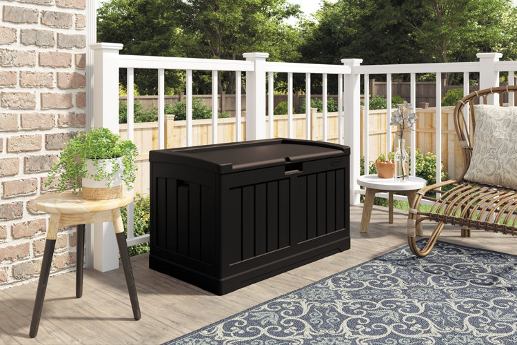 black plastic storage bench on a deck next to a wicker chair and blue area rug