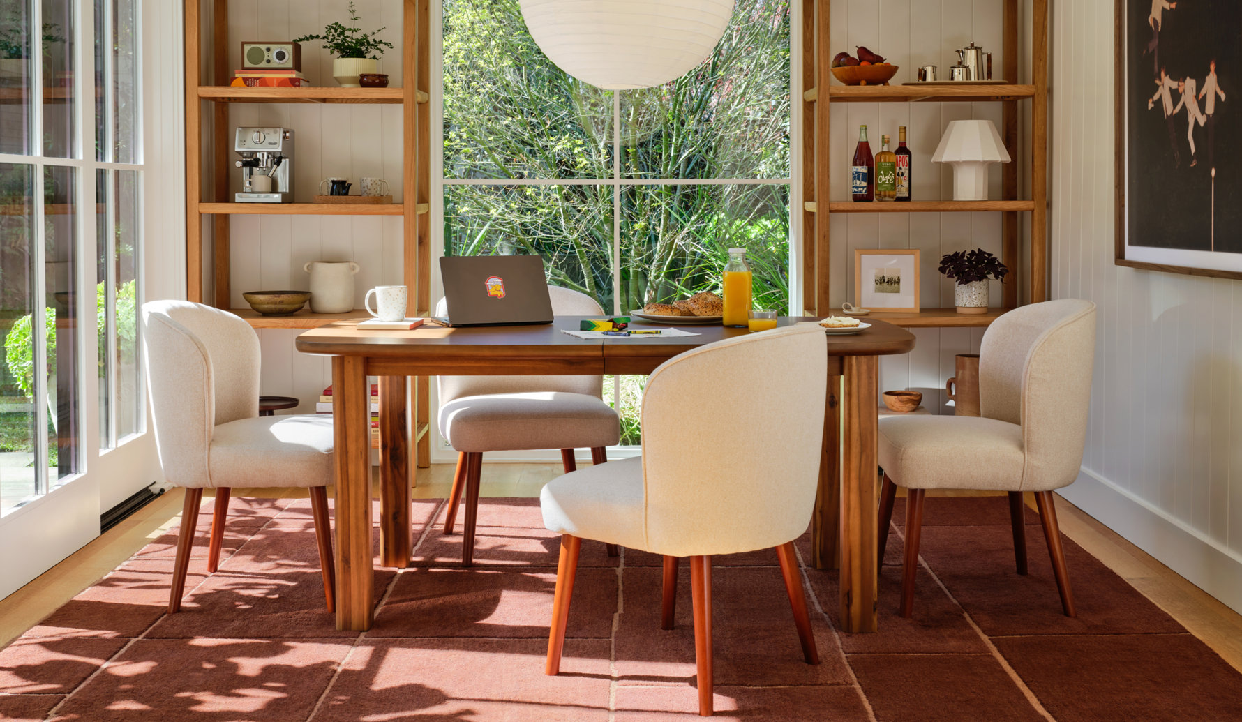 A dining room featuring a wooden table with four cream upholstered chairs set on a dark red tile rug in front of a large window with built-in wooden shelving on either side.