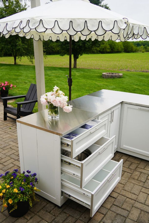 White outdoor kitchen island with a stainless steel countertop, drawers open, and a floral arrangement on top, set on a patio under a scalloped umbrella.