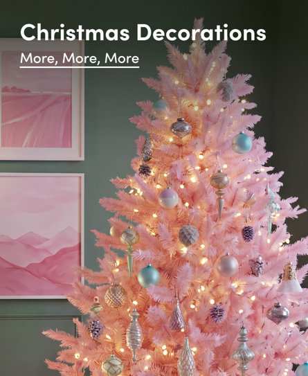Christmas Decorations: More, More, More. A vibrant pink artificial Christmas tree adorned with assorted baubles and warm fairy lights, set against a dark wall with two framed pink abstract landscape prints.
