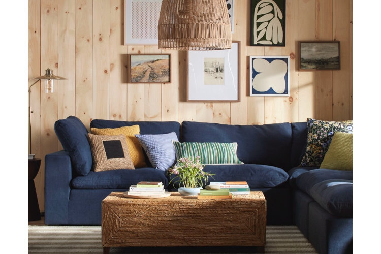 A cozy rustic living room featuring a large dark navy blue sectional sofa positioned against a light wood-paneled wall. The sofa is decorated with an eclectic mix of accent cushions, including a yellow knit pillow, a woven jute pillow, a blue gingham cushion, and a green striped lumbar pillow. In front of the sectional rests a rectangular woven wicker coffee table styled with stacks of books and a small flower arrangement. A gallery wall of framed botanical and abstract artwork hangs above the sofa, illuminated by a large woven pendant lamp hanging from the ceiling.