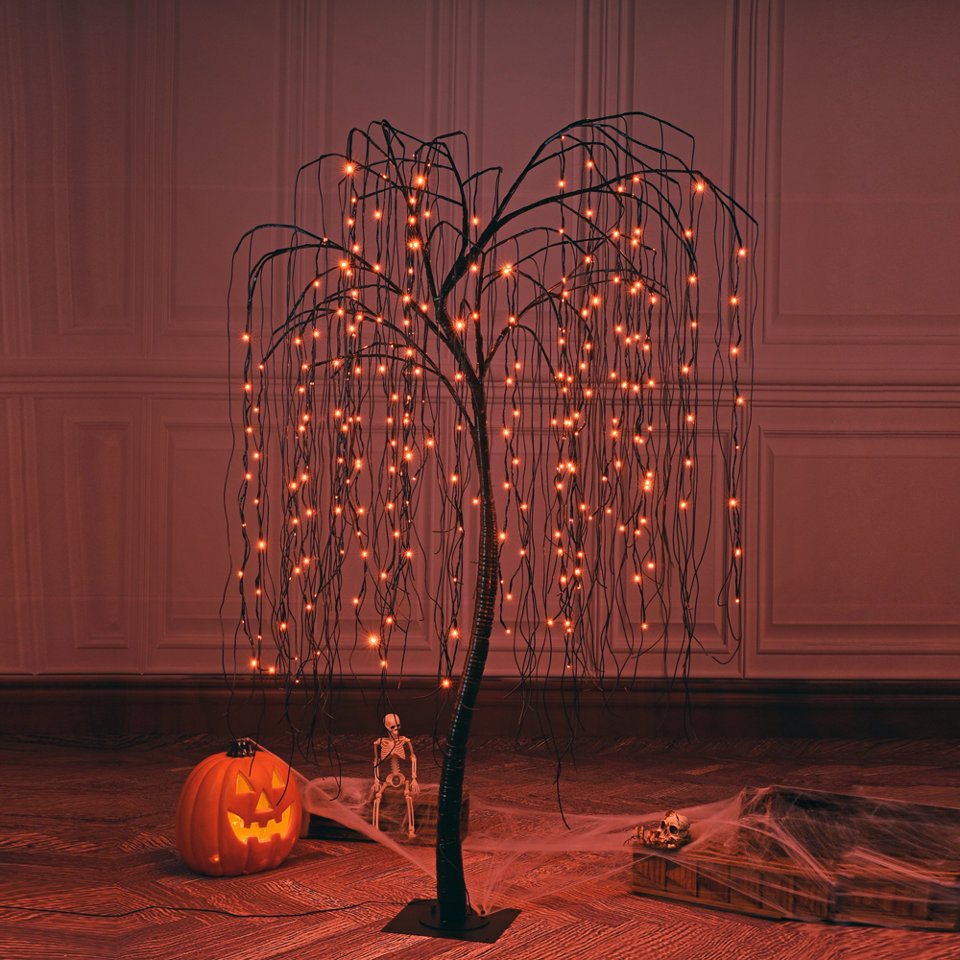 Pre-lit artificial weeping willow tree with orange LED fairy lights, placed on a floor with a jack-o'-lantern, small skeleton, and decorative spiderwebs for Halloween decor.