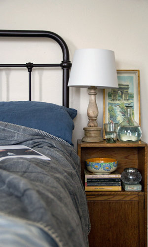 A bed with a cuddly grey throw blanket, wooden bedside lamp, water glasses and vase.