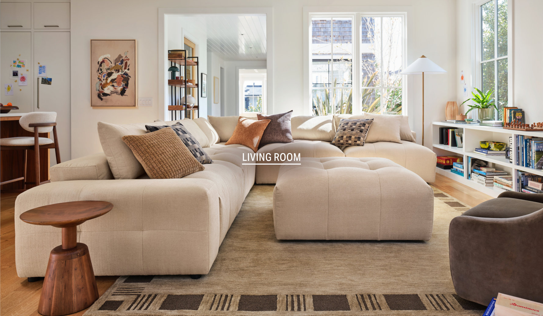 Bright, modern living room featuring a large, cream-colored modular sectional sofa, a tufted ottoman, and a cozy area rug, styled with various textured throw pillows.