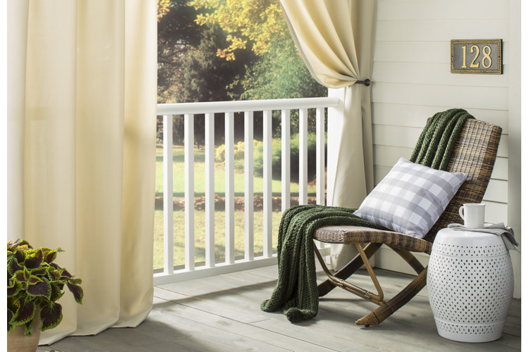 Cozy front porch seating area with wicker lounge chair, side table, and curtains, styled with pillows and a throw overlooking a garden.