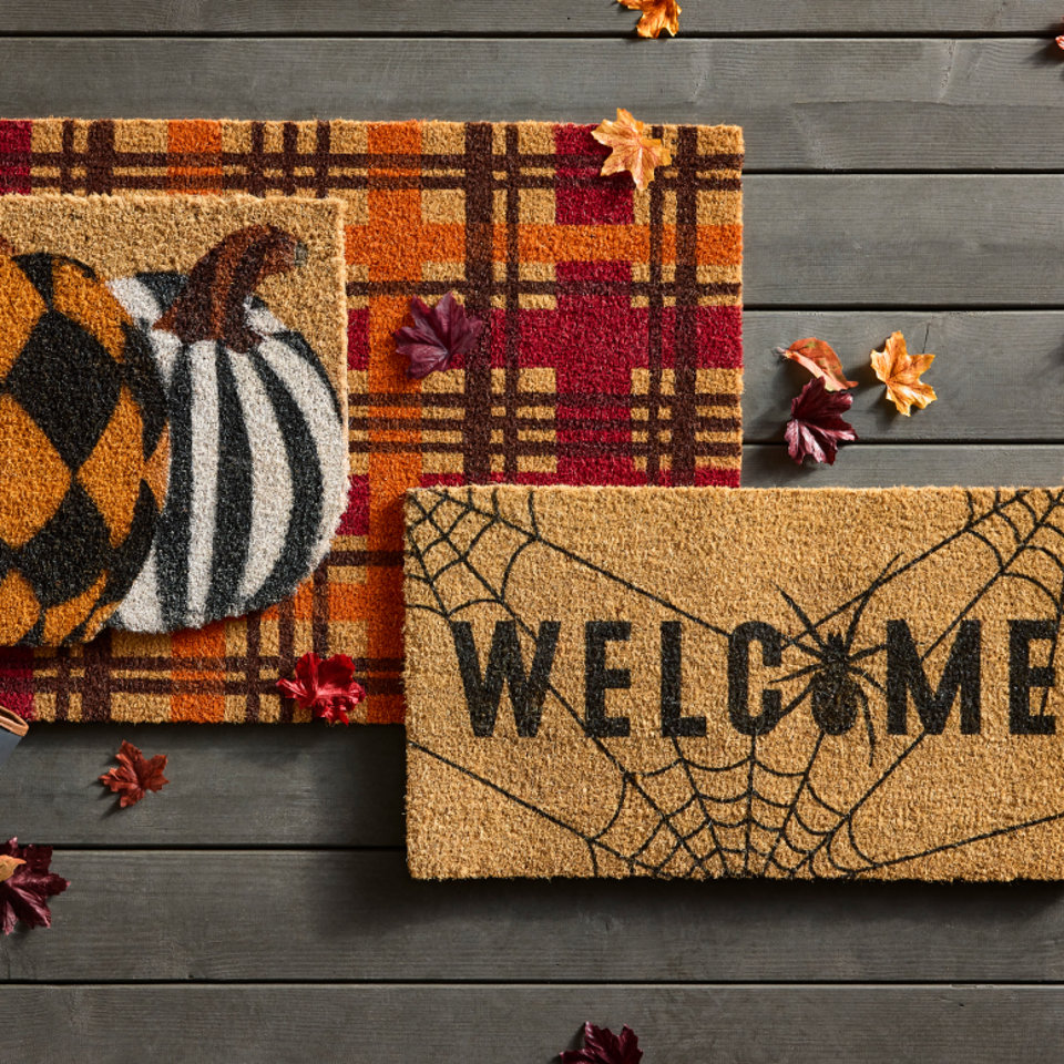 Three seasonal coir doormats on a wooden porch, featuring a plaid pattern, a decorative pumpkin, and a spiderweb design with the word Welcome.