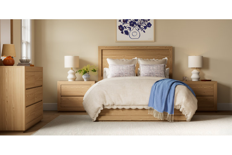 A serene modern bedroom featuring light oak furniture against cream-colored walls. Above the bed hangs framed art depicting blue flowers in a vase, while a matching light oak dresser sits on the left, and a cream area rug covers the hardwood floor.