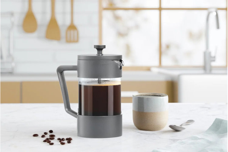 gray French press on a white marble countertop with a ceramic mug, cloth napkin, and scattered coffee beans around it