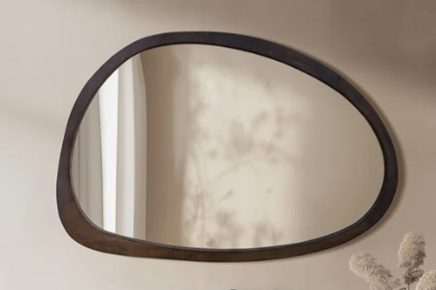 Asymmetrical wall mirror with a dark wood frame mounted on a neutral beige wall featuring organic leafy shadow accents.