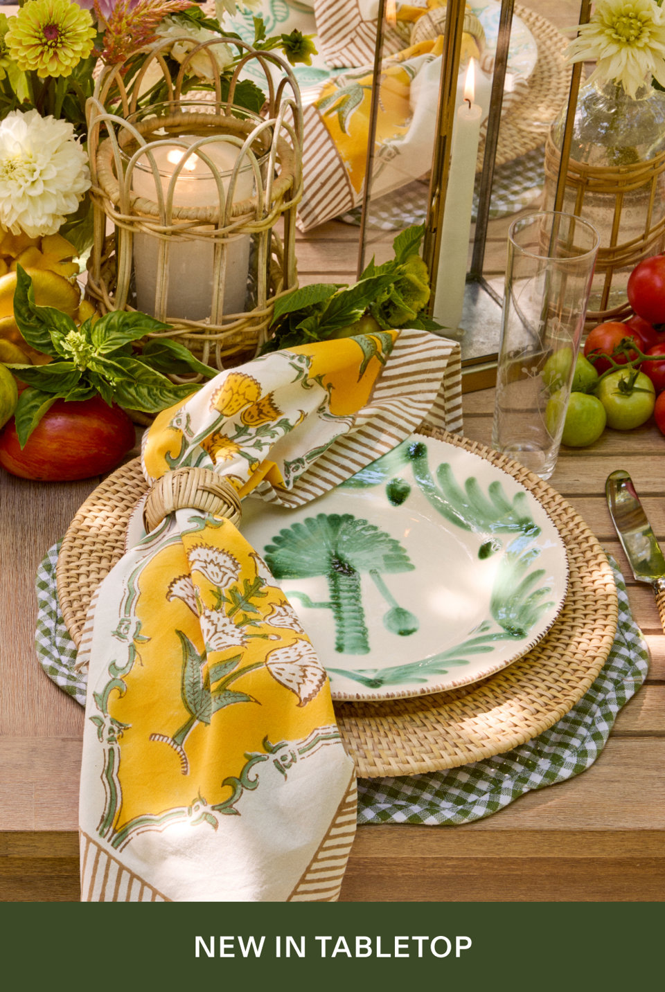 A summer table setting featuring hand-painted palm tree plates, yellow floral napkin with rattan ring, woven rattan chargers, green gingham placemat, rattan lanterns with candles, and fresh garden tomatoes. Text reads 'NEW IN TABLETOP'.