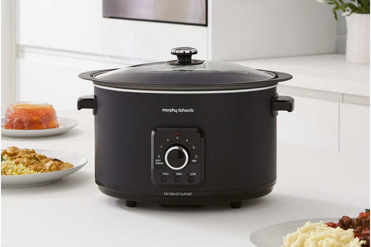 matte black slow cooker surrounded by plates of food on a white countertop