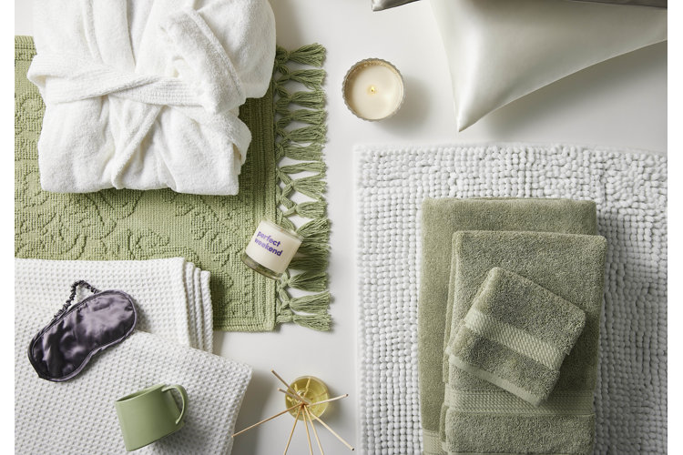 An assortment of white and green self-care items including a white bathrobe, candles, a green mug, and bath mats.