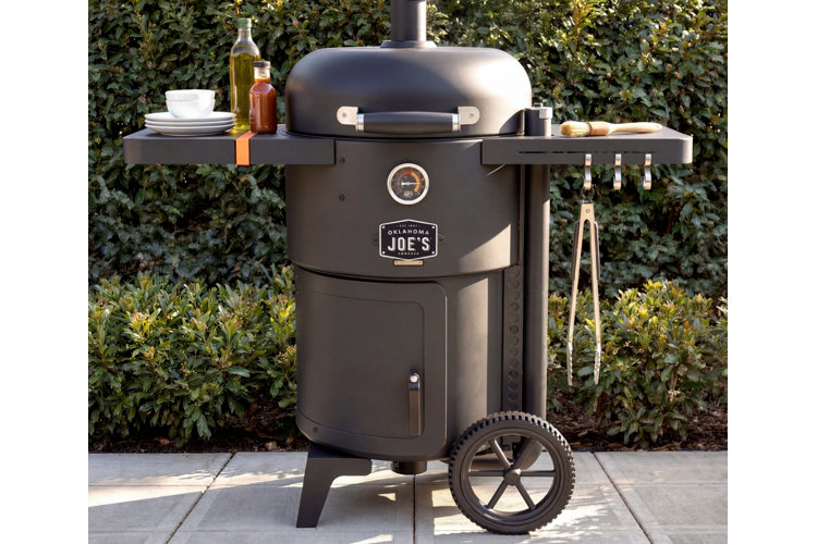 A matte black Oklahoma Joe's drum smoker and grill set outdoors on a stone patio in front of lush green foliage. The smoker features side shelves stocked with white plates, olive oil, sauce bottles, a basting brush, and hanging metal tongs, supported by large wagon-style wheels.