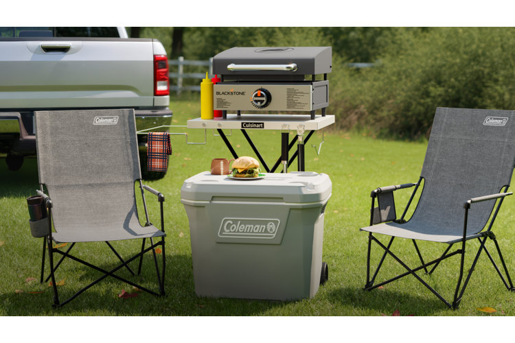 An outdoor tailgating setup on a grassy lawn with a silver pickup truck parked on the left. In the center, a portable Blackstone tabletop griddle rests on a white Cuisinart folding stand with mustard and ketchup bottles next to it. In front of the stand, a large gray Coleman wheeled cooler serves as a small table holding a burger on a plate and a mug. Two gray Coleman folding lawn chairs flank the cooler setup on either side.