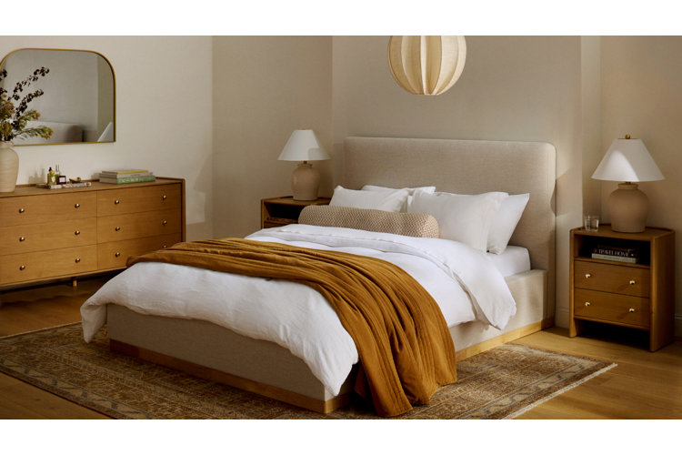 modern style bedroom with a white upholstered bed and headboard white bed set with a yellow throw blanket a brown and beige traditional patterned area rug, light wood colored dresser and matching nightstands on either side of the bed which is placed up against a wall