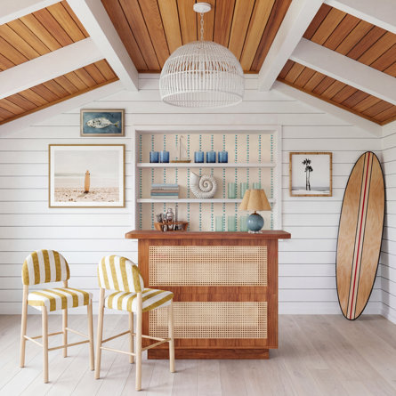 Bar de style côtelé en rotin avec deux tabourets rayés jaunes et blancs dans une pièce à plafond voûté en bois et lambris blanc.
