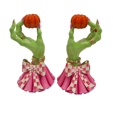 Two decorative candle holders shaped like green witch hands wearing gold rings and pink dresses with white checkered bows, each holding a small orange pumpkin.