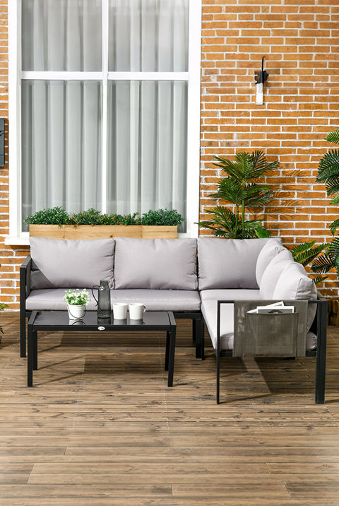 L-shaped black metal outdoor corner sofa with light grey cushions and a matching glass-top coffee table on a wooden patio against a brick wall.