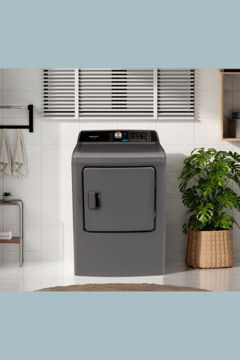 Gray Crosley top-load dryer centered in a bright, modern laundry room with white tiled walls, a large green plant in a woven basket, and window blinds.