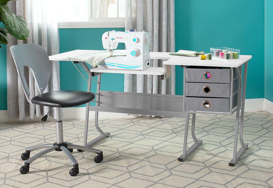 White sewing table with a drop-down platform, three gray storage drawers, and a matching rolling chair in a craft room.