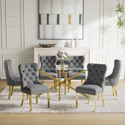 7-piece dining set with a glass-top rectangular table on a gold pedestal base and six gray tufted velvet upholstered chairs.