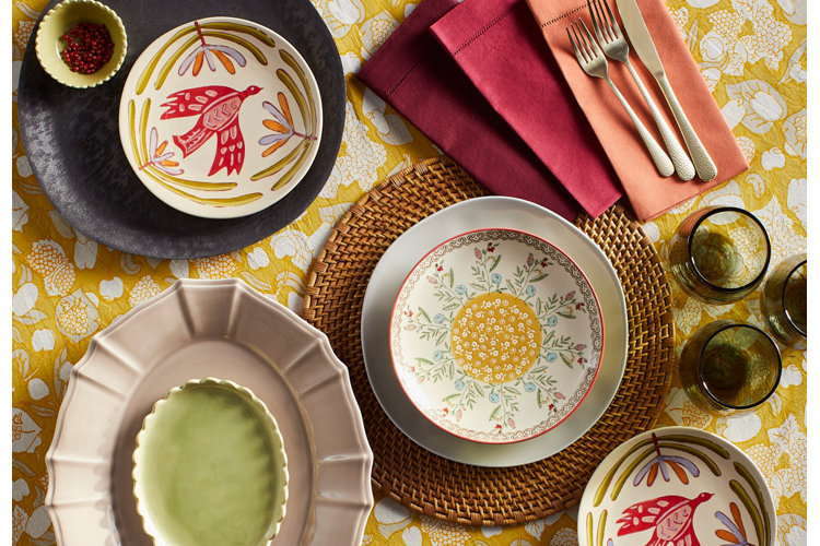 A vibrant, overhead shot of an eclectic table setting on a yellow and white patterned tablecloth. The assortment includes a woven rattan charger plate holding a porcelain white plate and a floral bowl with a yellow center, alongside smaller painted porcelain plates featuring a red painted bird design. A textured pink scalloped platter sits in the lower left next to a collection of stacked burgundy and peach napkins topped with silverware, with green glassware arranged on the right.