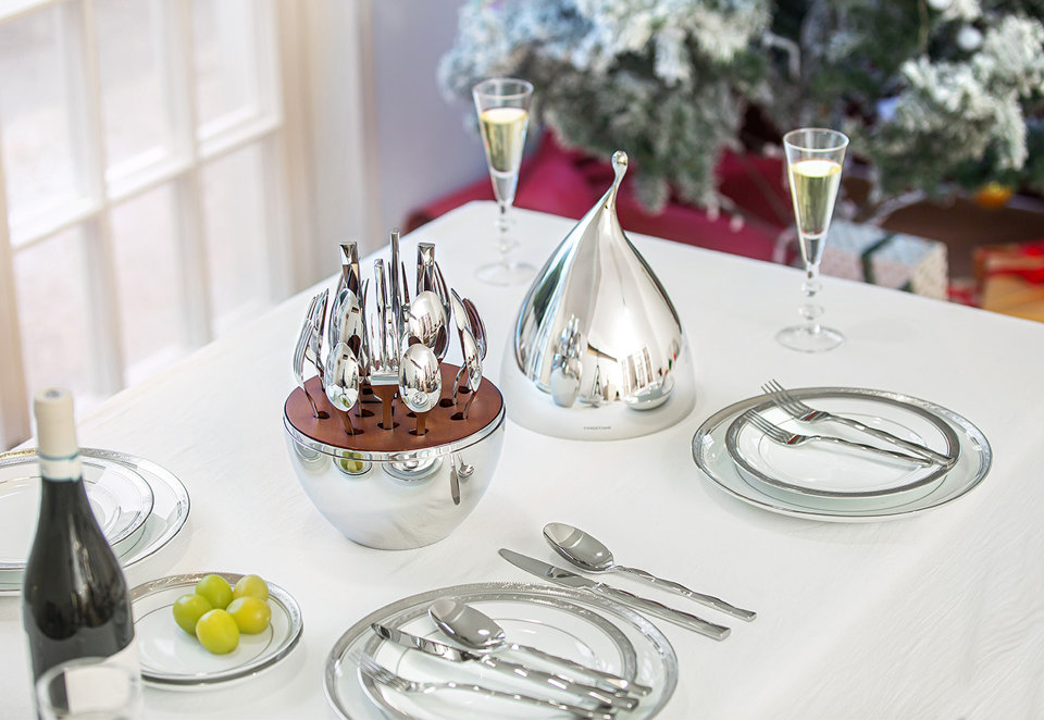 A formal dining table set with white plates, wine glasses, and a modern chrome tiered cutlery set in a wooden base with a matching silver dome cover.