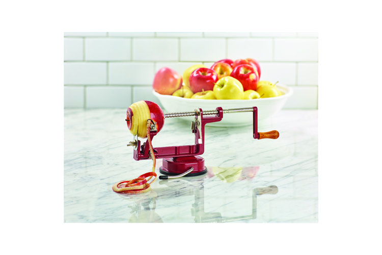 apple being peeled by a manual corer and peeler on a marble counter with a bowl of apples in the background