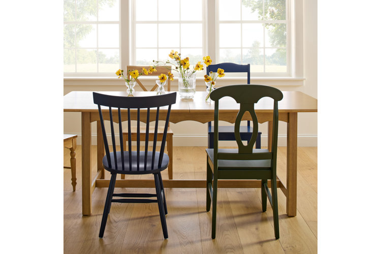 A bright, sunlit dining room featuring a long light oak table surrounded by a mismatched collection of dining chairs. The chairs include a tall ladder-back wood chair on the far left, a dark navy blue Windsor-style spindle chair in the foreground, a dark green splat-back chair on the right, and two more wood and blue chairs tucked behind the table. The table is minimally styled with clear glass vases holding cheery yellow cosmos flowers, set against a large multi-pane window that overlooks a bright outdoors.