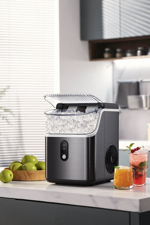 A dark, brushed-metal portable ice maker with its lid open, revealing a basket full of crushed ice, sitting on a kitchen counter next to a bowl of green apples and two glasses of colorful drinks.
