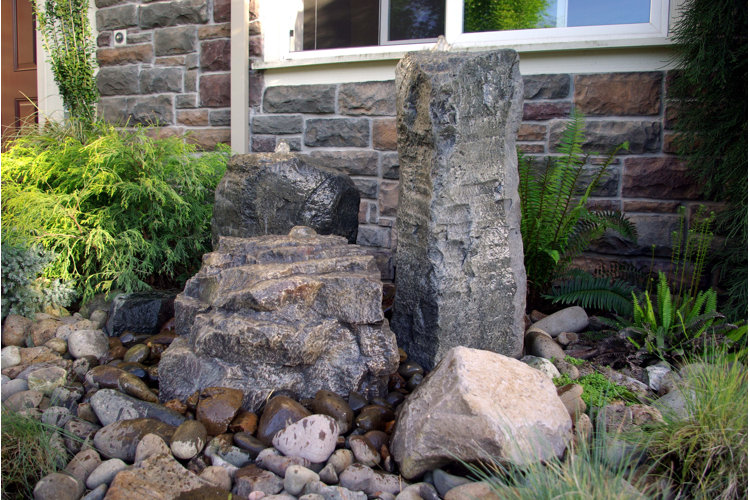 Low-maintenance xeriscape idea using large decorative boulders on top of a layout of smaller decorative rocks and green drought-resistant plants mixed in infront of a natural stone wall of the outside of a house.