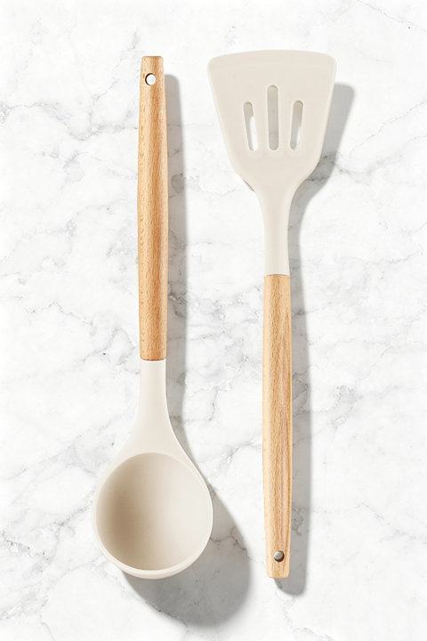 Cream silicone ladle and slotted turner set with light wood handles arranged on a white marble surface.