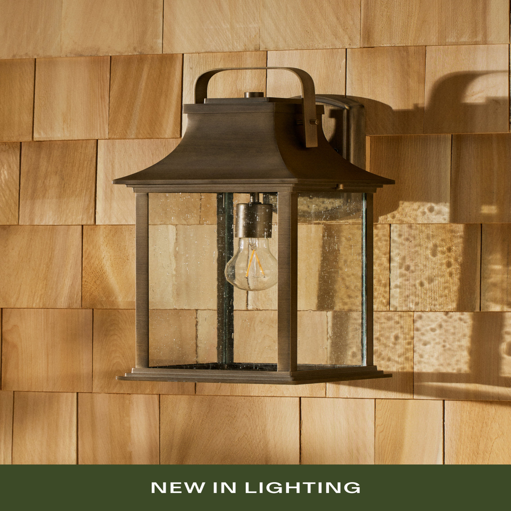 A bronze outdoor wall lantern with seeded glass panels and an Edison-style bulb, mounted on natural cedar shake siding. Text reads 'NEW IN LIGHTING'.