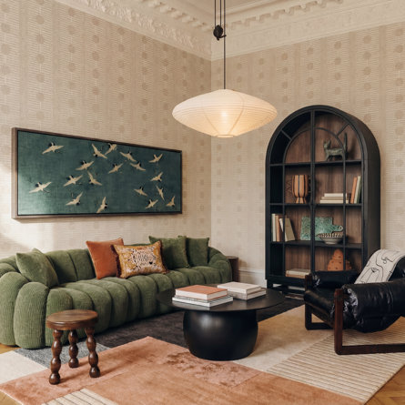 Vintage maximalist living room featuring a green curved sofa, crane artwork on the wall, an arched bookshelf, leather accent chair, textured wallpaper, and layered area rugs