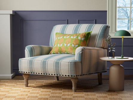 A striped upholstered armchair in cream and light blue with nailhead trim, wooden legs, and a decorative pillow, next to a small side table.