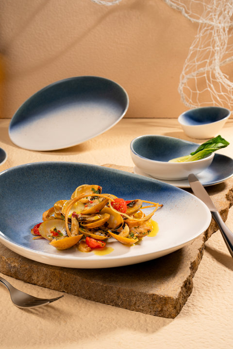 Clam pasta served in a blue and white ombre ceramic bowl with matching dinnerware on a stone table setting.