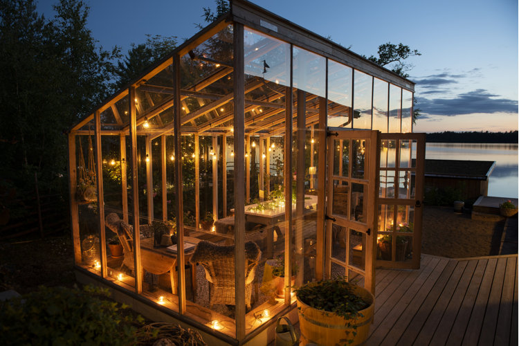 A glass solarium by a lake at dusk with string lights inside and around it
