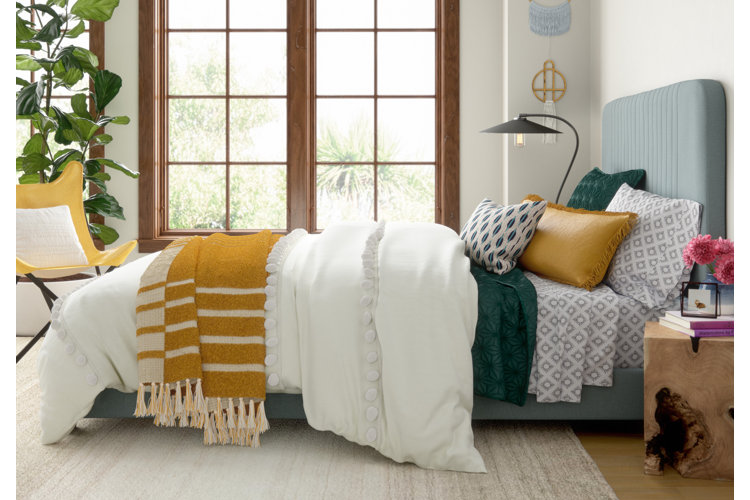 An upholstered bed with layers of white, yellow, and blue bedding with stripes, geometric patterns, and embroidered designs.