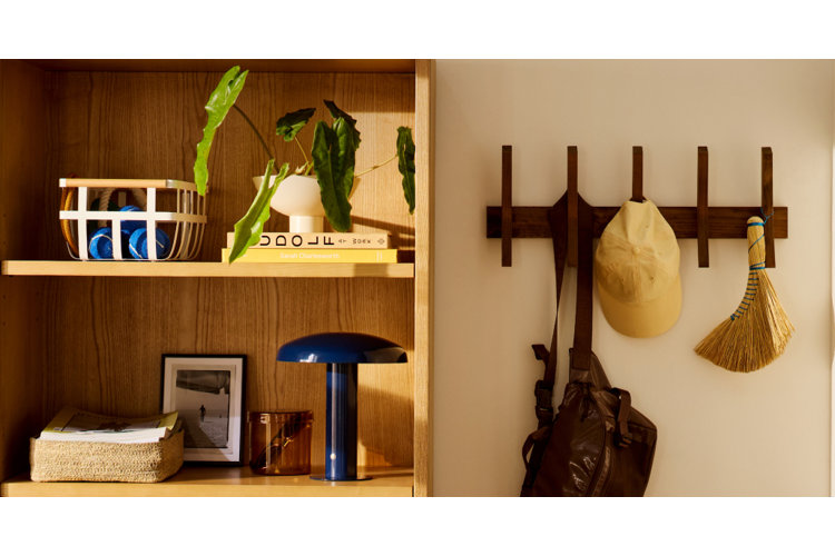 A brightly lit entryway scene featuring an open wooden shelving unit on the left and a wall-mounted wooden coat rack on the right. The top shelf holds a wire basket with blue tennis balls and a potted green plant sitting on books. The bottom shelf displays a woven basket filled with papers, a small framed picture, an amber glass cup, and a sleek blue mushroom-shaped table lamp. Hanging from the vertical hooks of the coat rack are a dark brown backpack, a beige baseball cap, and a small handheld straw broom wrapped with blue thread.