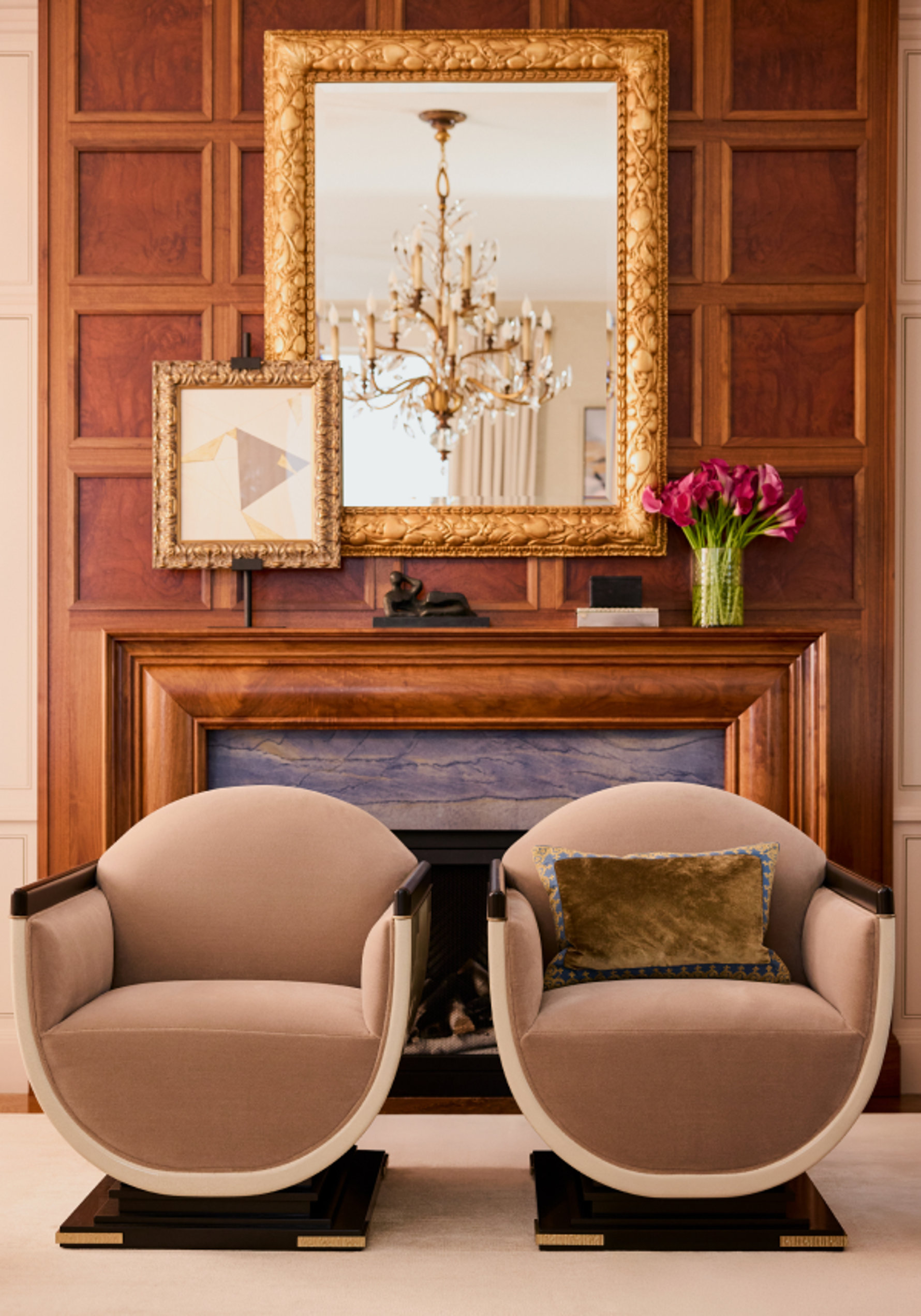 Two round armchairs in front of a fireplace and gold mirror.