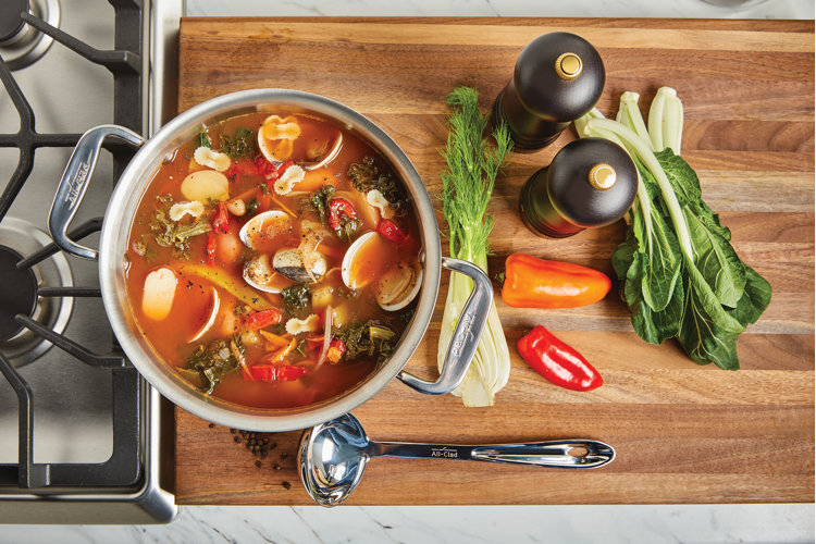 stainless steel soup ladle on a wooden cutting board with the ingredients surrounding a seafood stew in a stock pot.